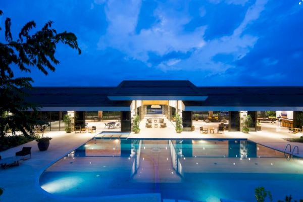 A modern building with large glass windows overlooks a pool under a dusky blue sky, with lights illuminating the area.