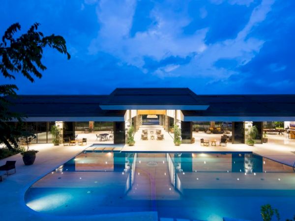 A modern building with large glass windows overlooks a pool under a dusky blue sky, with lights illuminating the area.