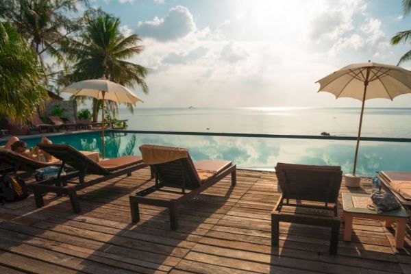 Poolside scene with sun loungers and umbrellas on a wooden deck, overlooking a serene body of water under a partly cloudy sky.