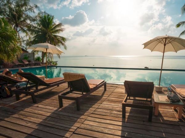 Poolside scene with sun loungers and umbrellas on a wooden deck, overlooking a serene body of water under a partly cloudy sky.