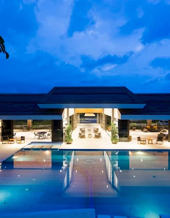 A luxurious modern building with a large illuminated swimming pool and open indoor seating area under a dramatic evening sky.
