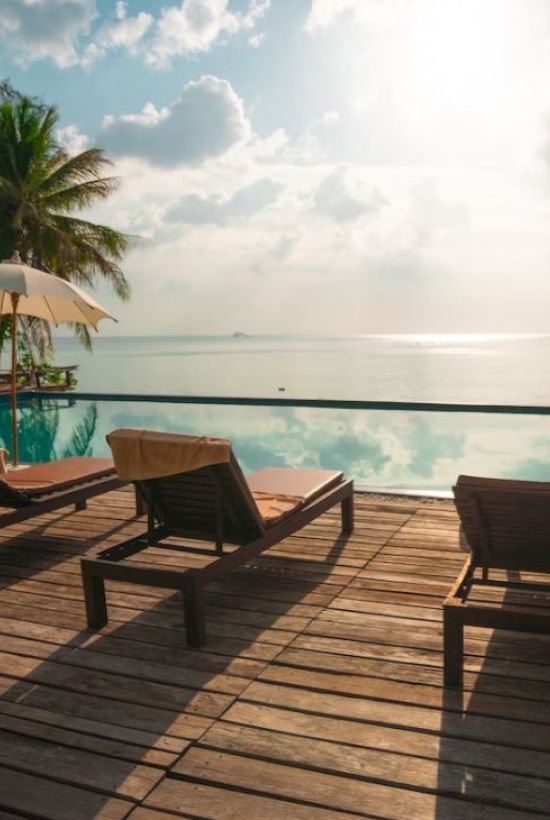 Lounge chairs and umbrellas by a pool, overlooking the ocean with palm trees and a partly cloudy sky in the background.