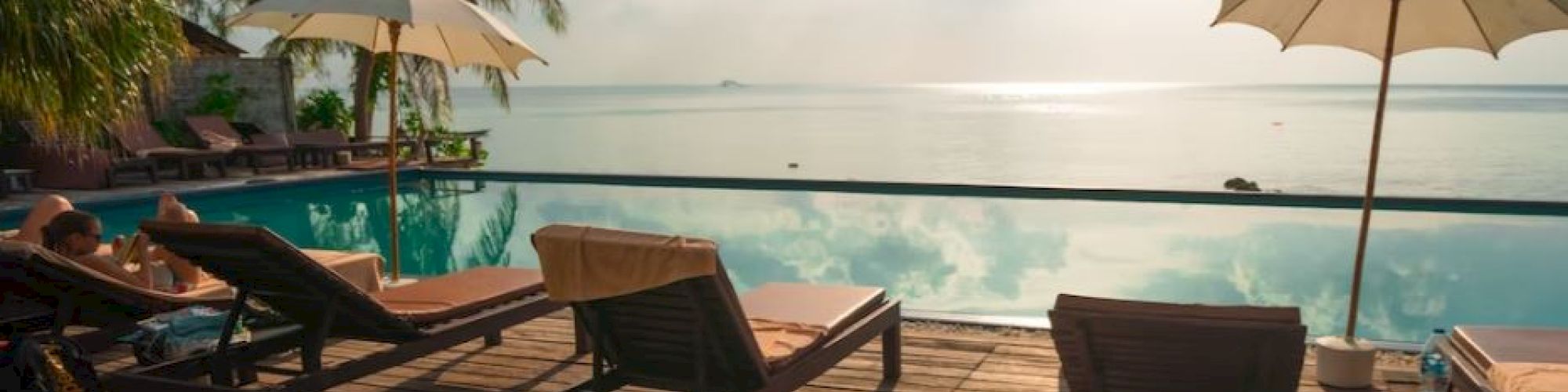 A serene poolside scene with lounge chairs, umbrellas, and palm trees overlooking a calm sea under a cloudy sky.
