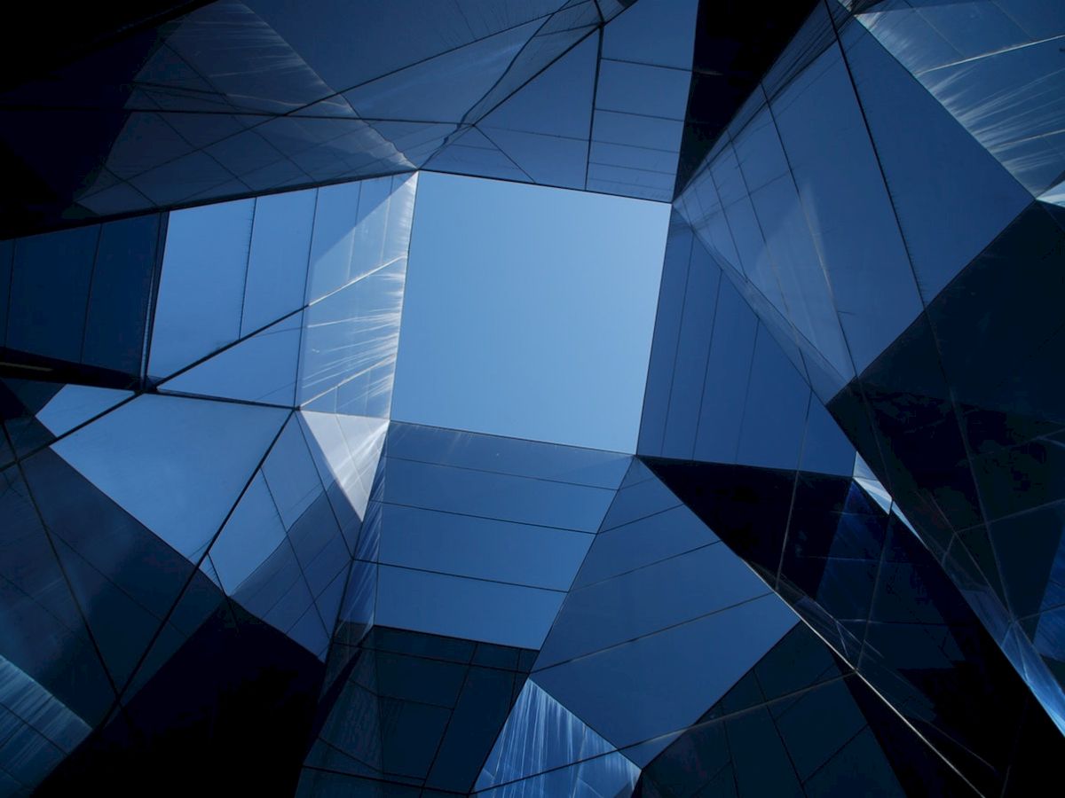 The image shows a geometric pattern of blue glass and metal panels forming an abstract architectural structure with a clear sky above.