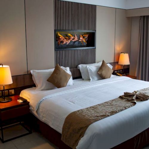 A modern hotel room features a large bed with brown accents, two bedside tables with lamps, and a window with curtains.