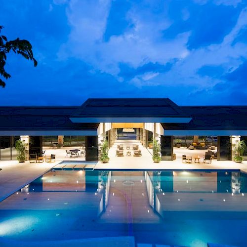 A luxurious modern building with an illuminated pool at night, surrounded by trees and open seating areas under a vibrant blue sky.