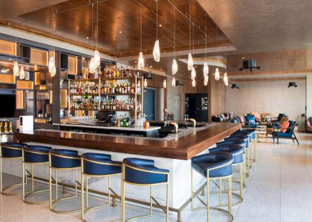 The image shows a modern bar with a wooden counter, blue chairs, pendant lights, and a fully stocked shelf of bottles in the background.