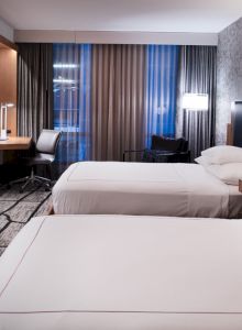 A modern hotel room with two beds, a TV, desk, and large window with curtains, featuring stylish decor and ambient lighting.
