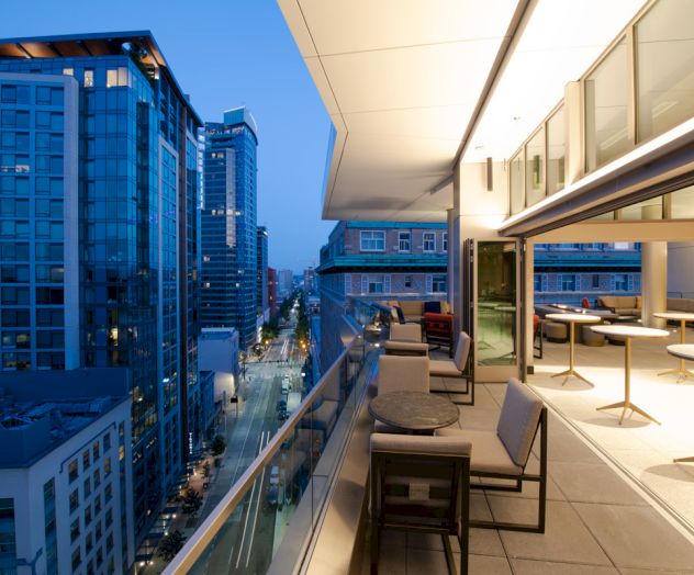 A modern cityscape at dusk shows high-rise buildings, a street below, and a sleek balcony with seating and tables under warm lights.