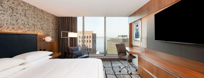 A modern hotel room features a large bed, wall-mounted TV, desk with chair, and window with a city view, offering a cozy ambiance.