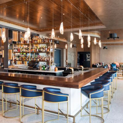 A modern bar with blue chairs, pendant lights, and a stocked counter sits centrally, surrounded by a stylish lounge area with seating.