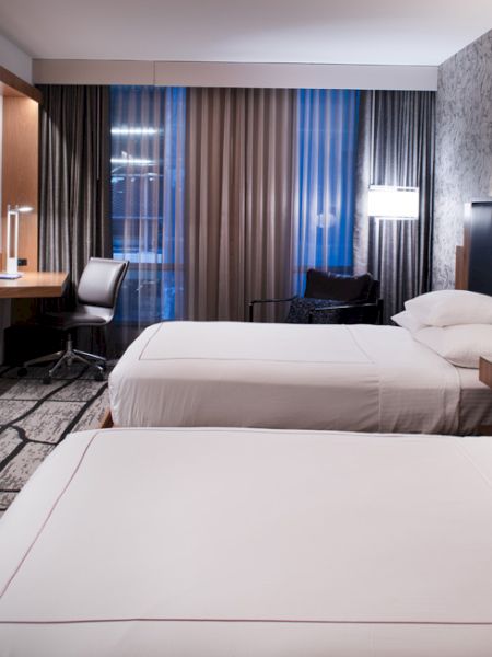 The image shows a modern hotel room with two beds, a flat-screen TV, a desk with a chair, and ambient lighting.