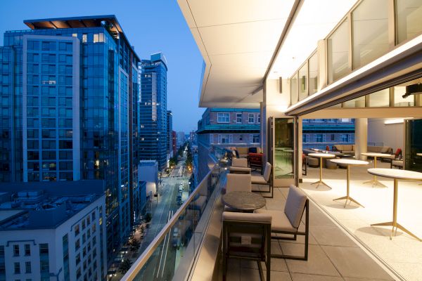 The image shows a modern rooftop terrace in an urban setting with cityscape views, featuring tables and chairs in the foreground.
