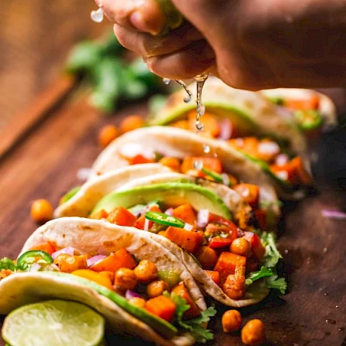 A hand squeezes lime over tacos filled with chickpeas, avocado, and veggies on a wooden board, garnished with cilantro and lime slices.