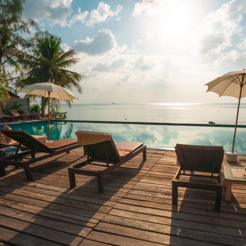 Beachfront scene with lounge chairs, parasols, and a pool overlooking the ocean under a bright sky with fluffy clouds.