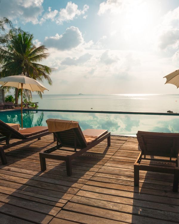 Beachfront scene with lounge chairs, parasols, and a pool overlooking the ocean under a bright sky with fluffy clouds.