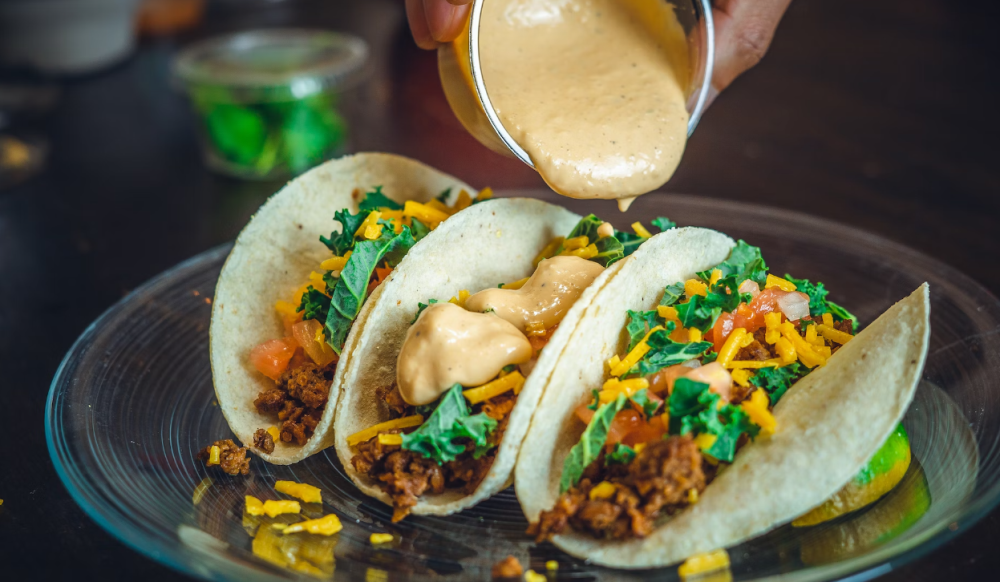 Three tacos with ground meat, cheese, lettuce, and sauce being poured from a cup, served on a clear plate with a lime wedge.