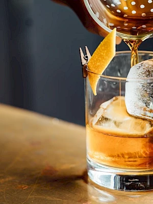 A cocktail is being poured into a glass with ice and a citrus peel garnish on a table.