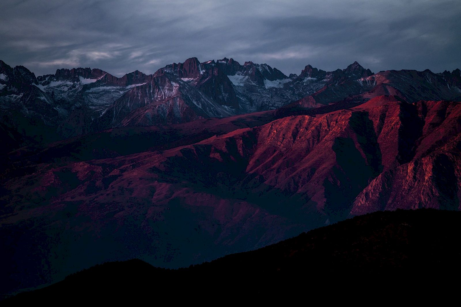 A mountain range under a moody sky, with peaks and ridges illuminated by a red-orange glow, possibly at sunrise or sunset.