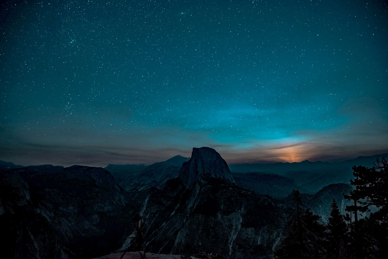 A stunning night sky full of stars above a silhouetted mountain landscape, with a glowing moon or setting sun on the horizon.