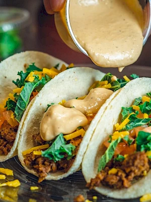 Three tacos on a plate filled with meat, cheese, lettuce, and sauce being poured over them.