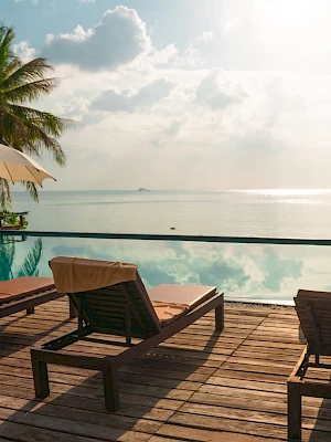 Poolside view with lounge chairs, umbrellas, and palm trees overlooking a serene body of water under a partly cloudy sky.