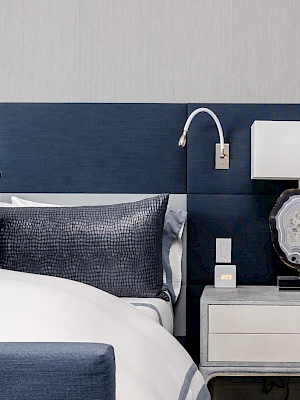 A modern bedroom features a blue upholstered bed, matching pillows, a nightstand with a lamp, and a wall-mounted reading light.