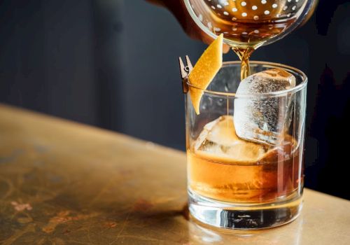 A close-up of a cocktail being poured into a glass with ice and a lemon twist garnish on a bartop surface.