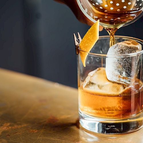 A close-up of a cocktail being poured into a glass with ice and a lemon twist garnish on a bartop surface.