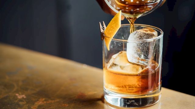 A close-up of a cocktail being poured into a glass with ice and a lemon twist garnish on a bartop surface.