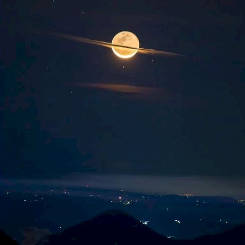 A glowing full moon with clouds resembling Saturn's rings, over a landscape with scattered city lights in the distance.