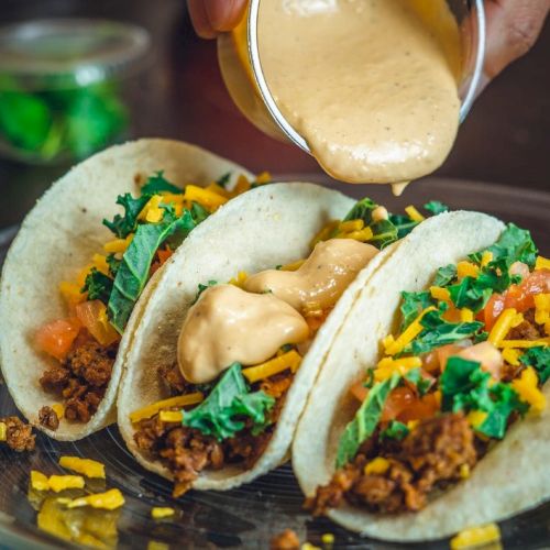 Three tacos filled with ground meat, cheese, lettuce, and tomato, topped with creamy sauce being poured over them.