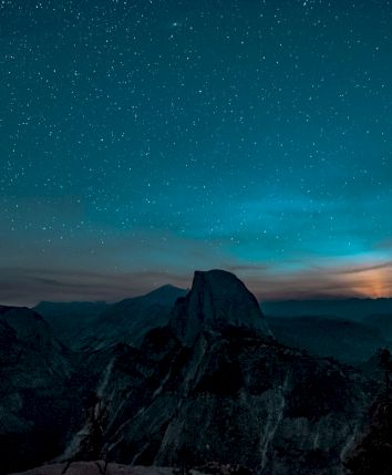 A night sky filled with stars above a mountainous landscape, with a visible horizon glow and silhouetted trees framing the scene.