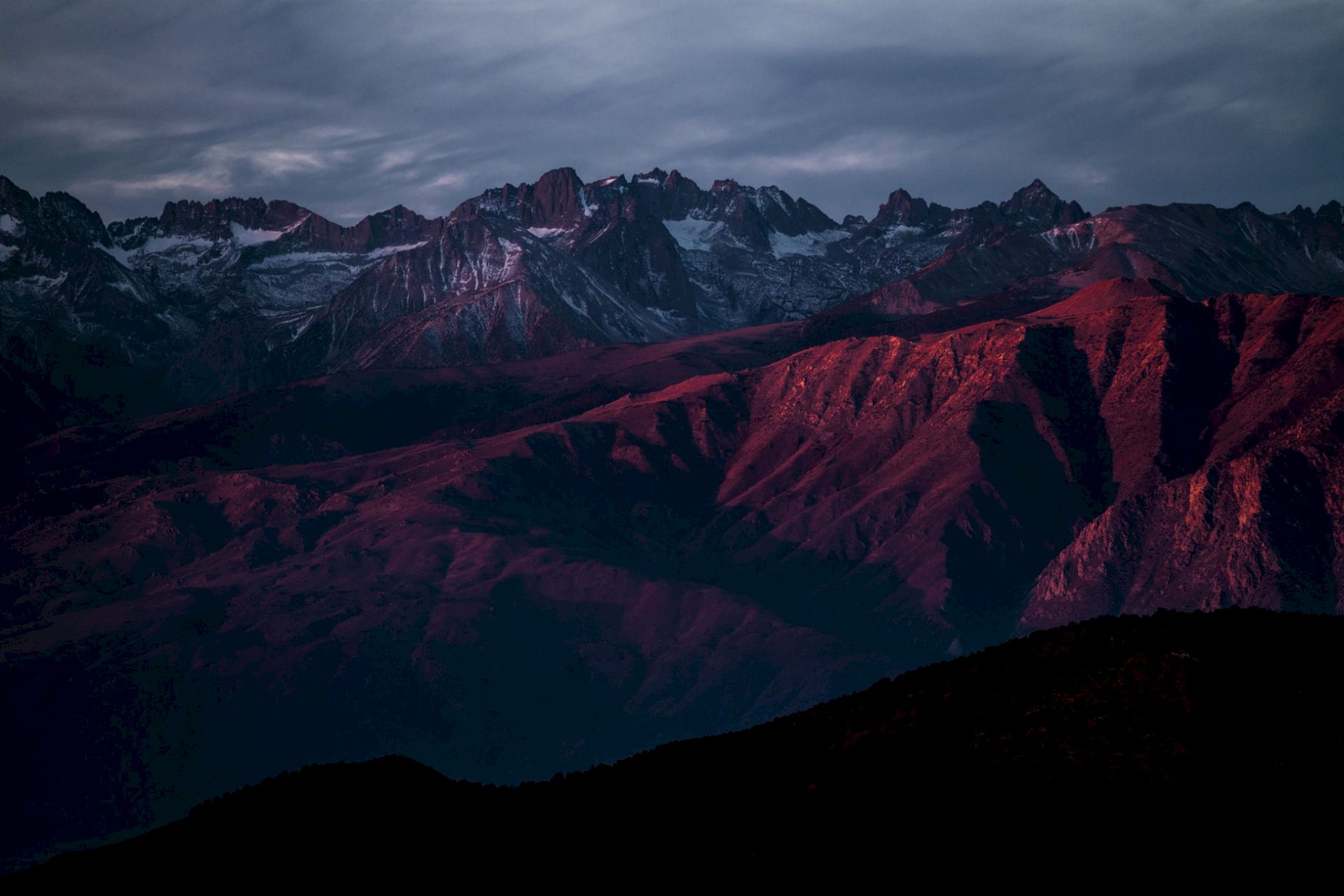 Majestic mountains with rugged peaks under a dramatic sky, bathed in red sunlight, create a stunning and serene landscape.