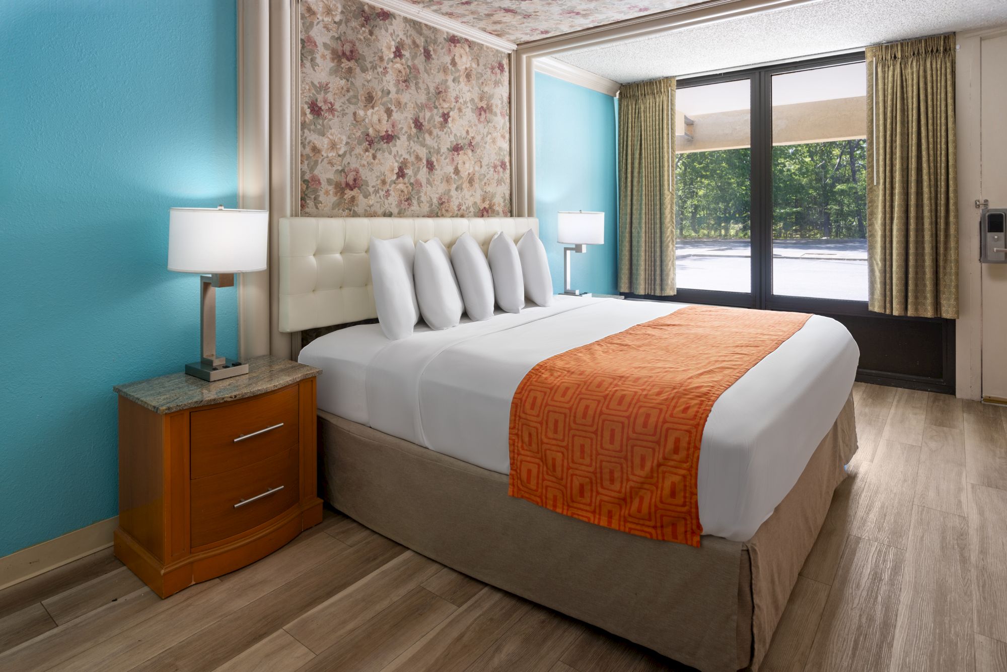 A cozy hotel room with a neatly made bed, orange throw, bedside tables, lamps, and a large window with curtains showing a greenery view.