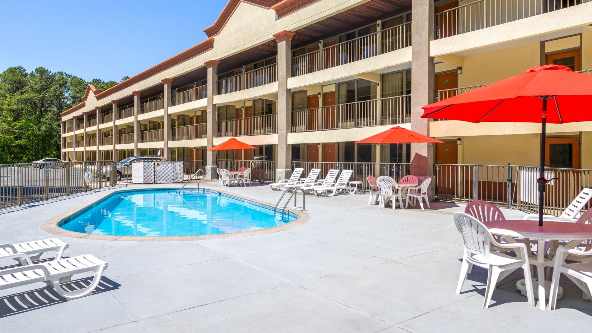 An outdoor pool area with sun loungers, red umbrellas, and tables. The setting is beside a multi-story building with balconies.