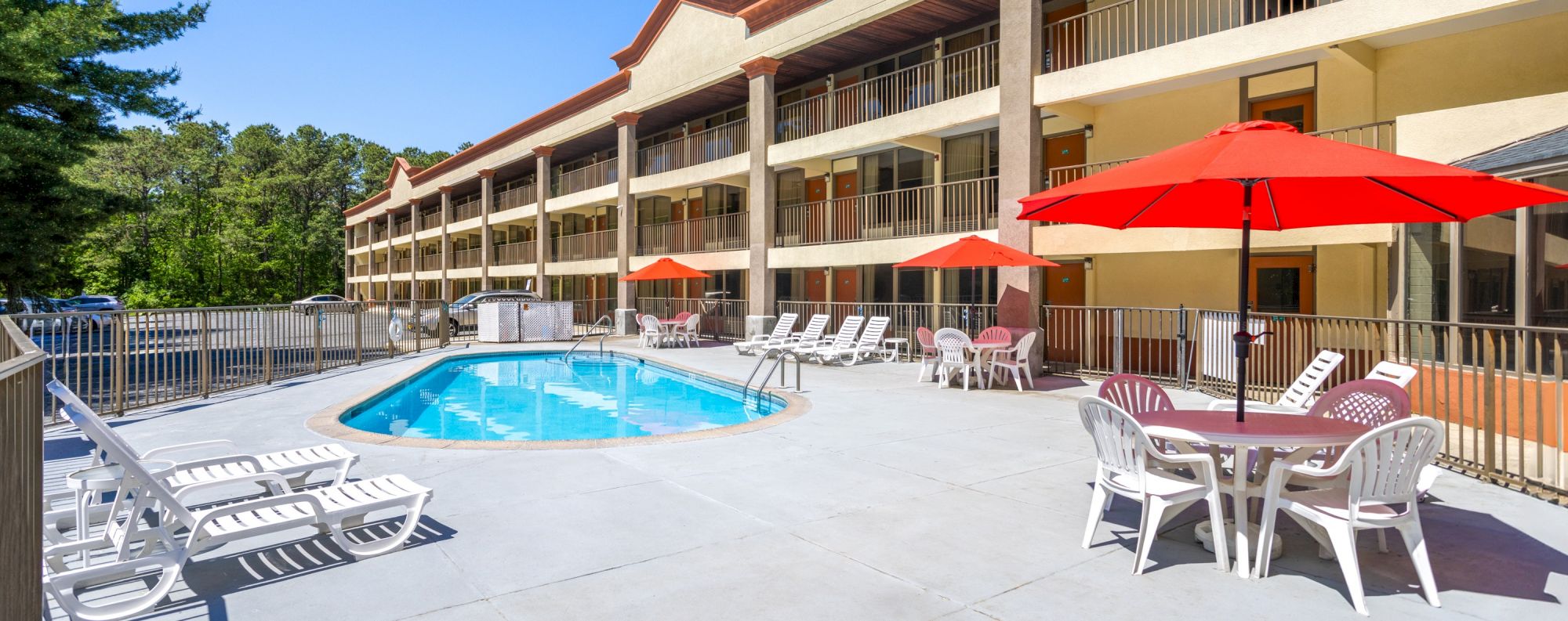An outdoor pool area with sun loungers, red umbrellas, and tables. The setting is beside a multi-story building with balconies.