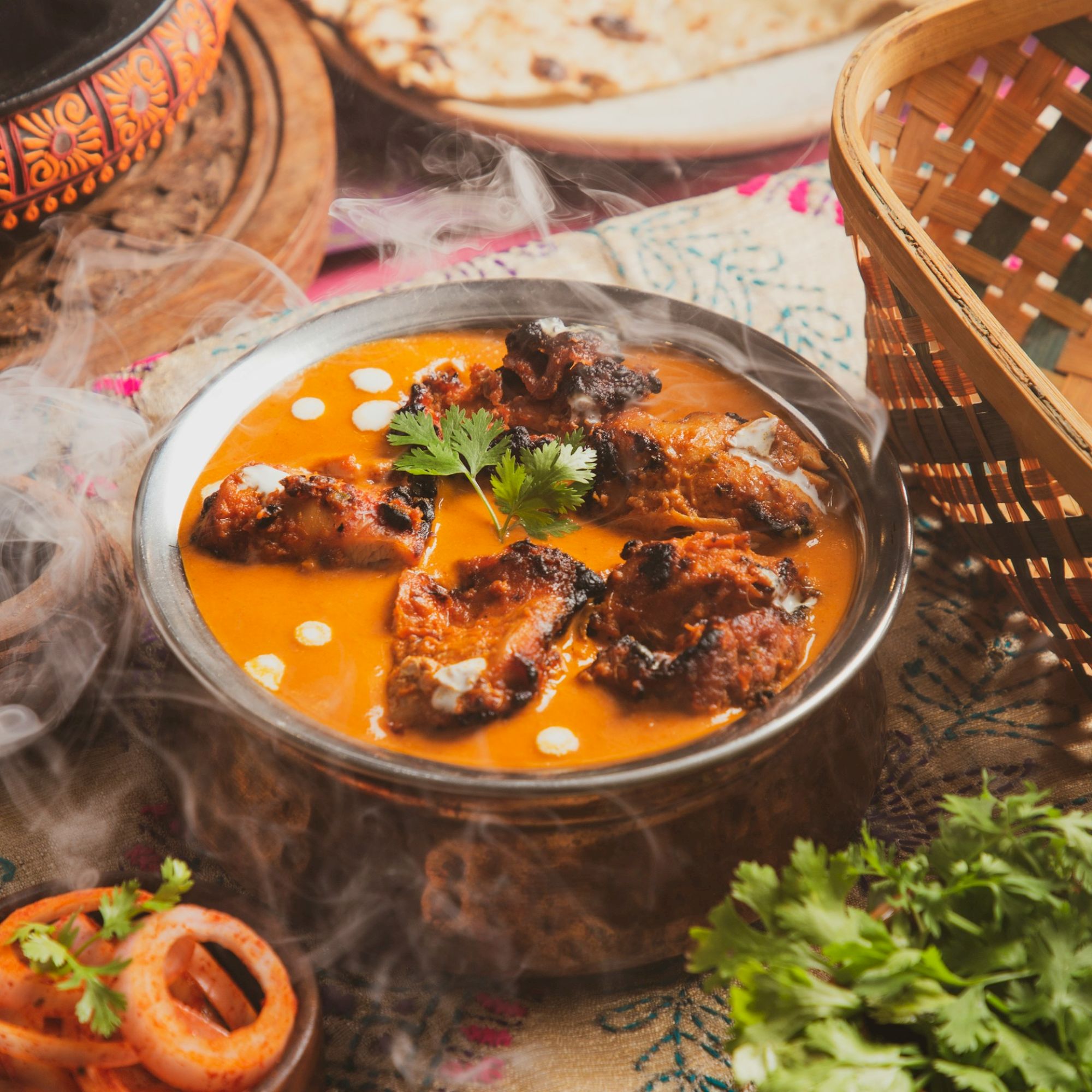 A steaming bowl of creamy curry garnished with herbs, served with naan and accompanied by fresh coriander and onion slices.