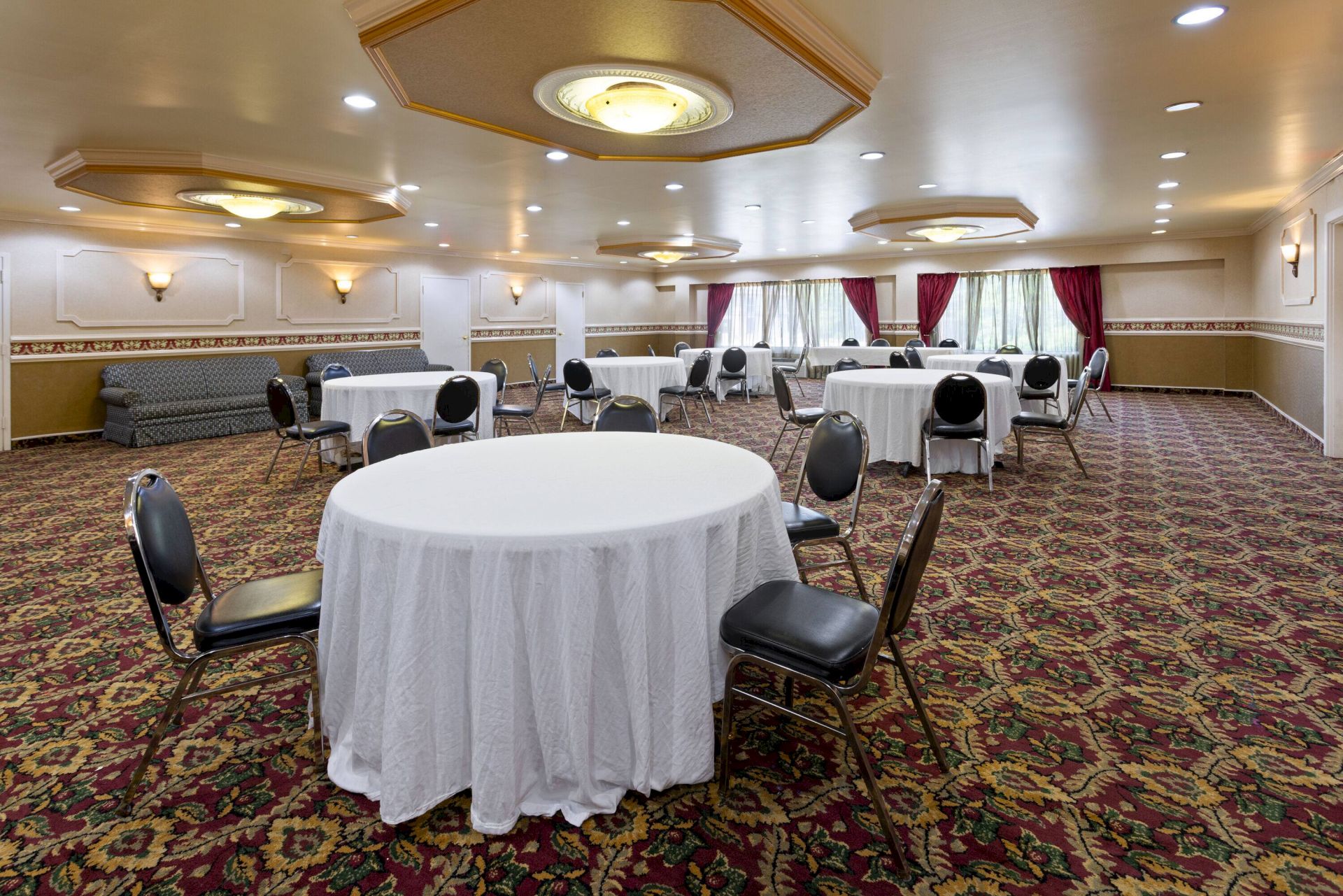 A banquet room with round tables covered in white cloth, black chairs, patterned carpet, and wall lights, ready for an event.