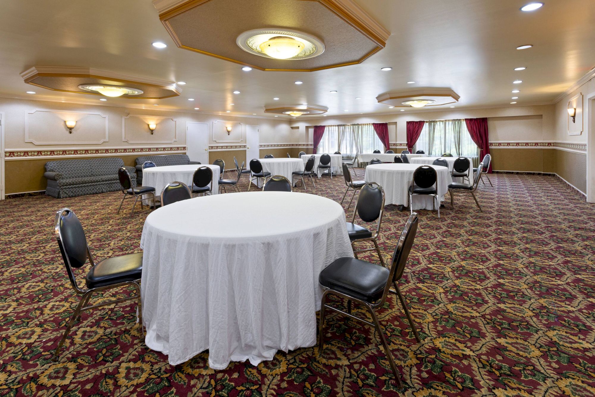 A banquet room with round tables covered in white cloth, black chairs, patterned carpet, and wall lights, ready for an event.