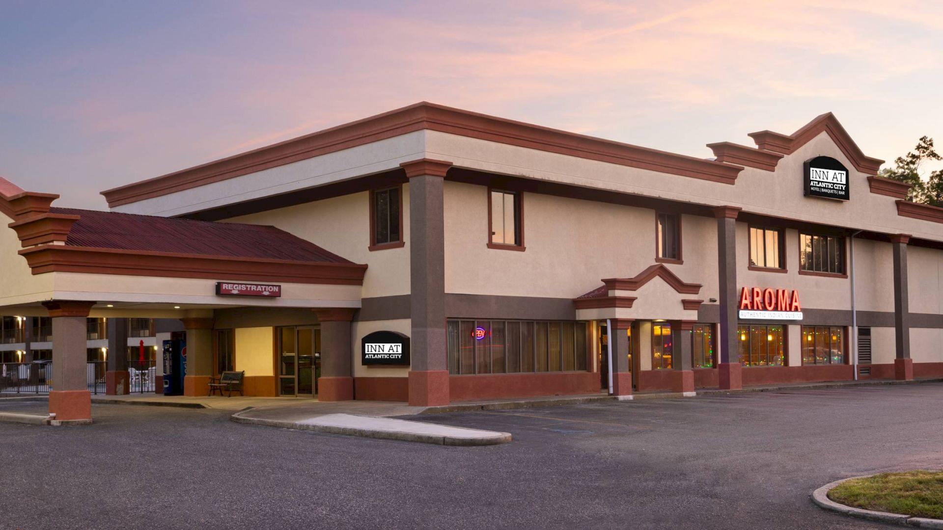 A two-story beige commercial building with red trim houses several businesses, including a sign reading &ldquo;AROMA&rdquo; and a large parking lot.