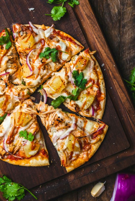 A pizza topped with chicken, pineapple, onions, cilantro, and cheese on a wooden board with fresh garnishes nearby.
