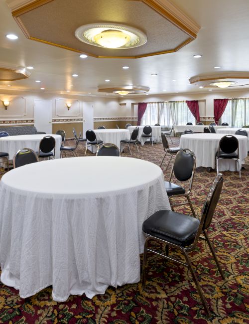 A banquet hall with round tables covered in white cloth and surrounded by chairs on a patterned carpet. Decor includes curtains and wall sconces.