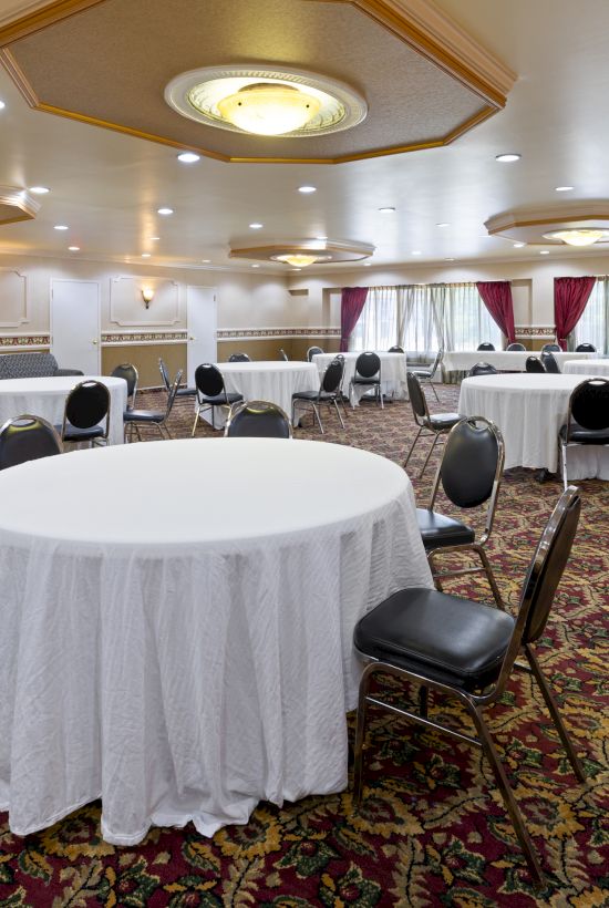 A banquet hall with round tables covered in white cloth and surrounded by chairs on a patterned carpet. Decor includes curtains and wall sconces.
