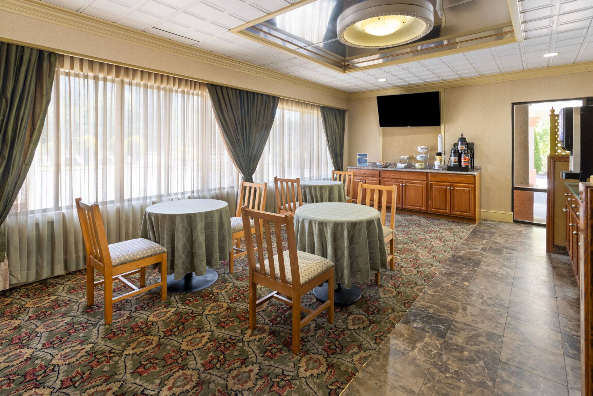 The image shows a dining area with tables, chairs, a patterned carpet, large windows, and a counter with a TV and coffee station.