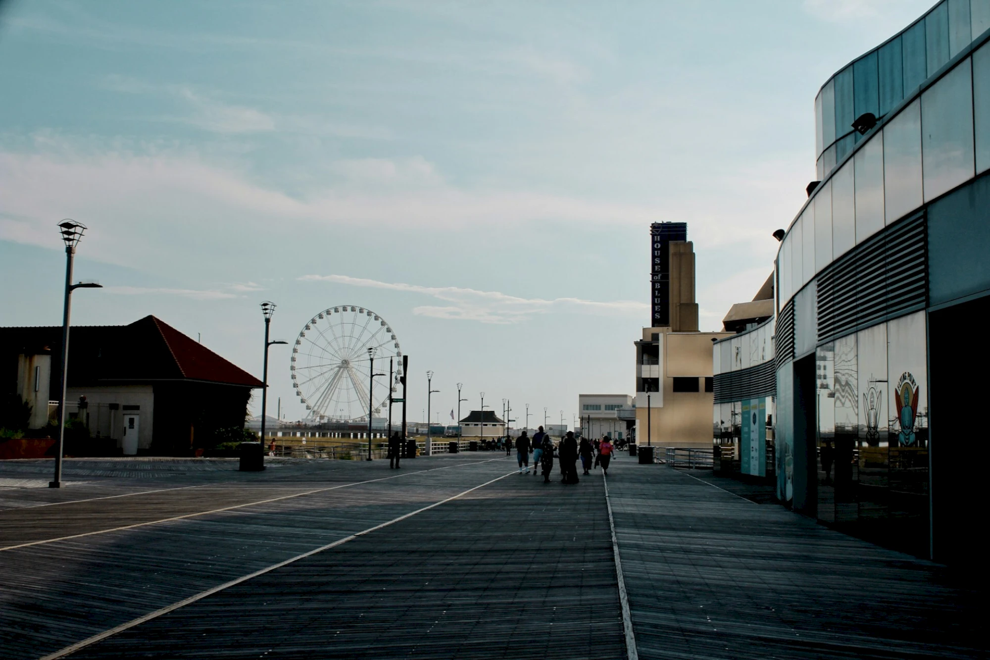 An outdoor plaza with people walking past a Ferris wheel and tall &ldquo;HOUSE&rdquo; tower beside modern buildings under a cloudy sky.