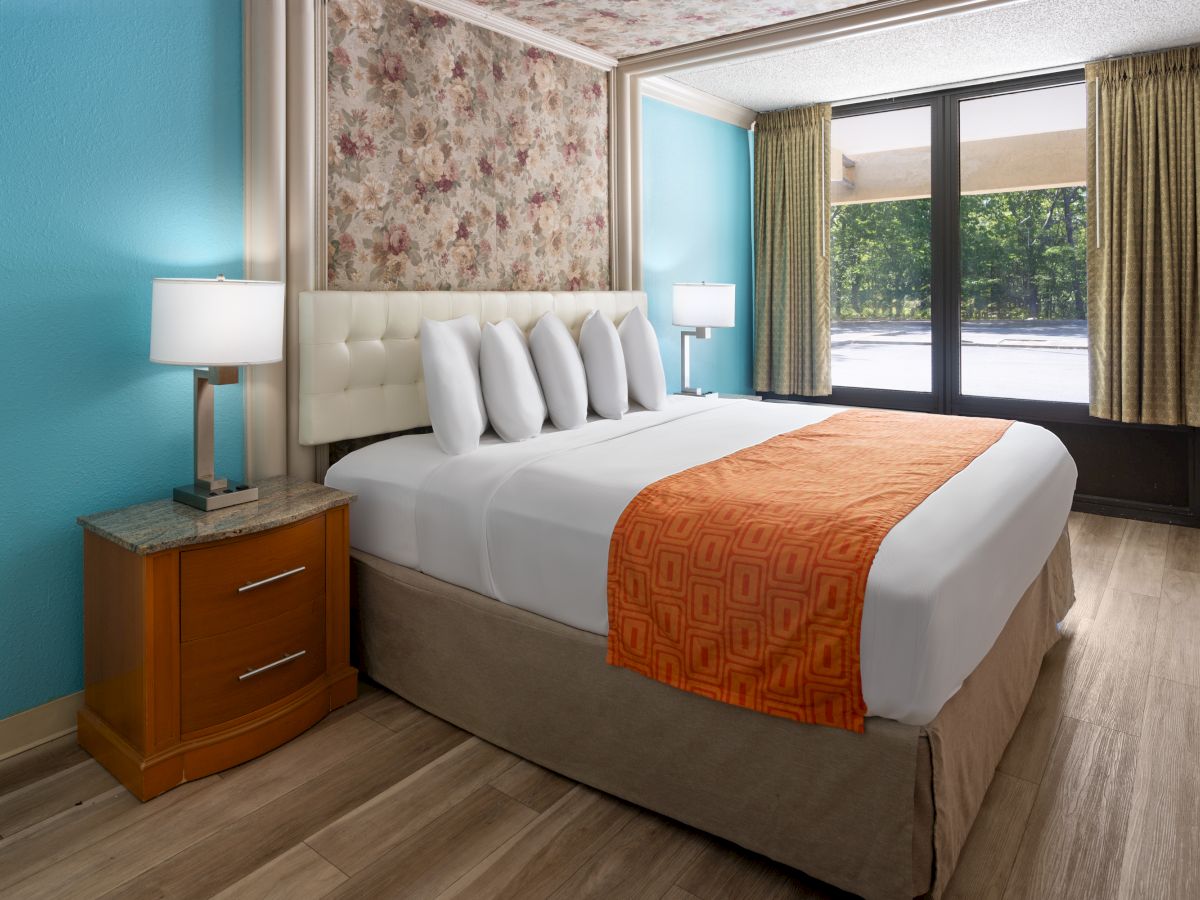 A hotel room with a bed, orange bed runner, nightstand with lamp, floral accent wall, blue wall, large window with curtains.