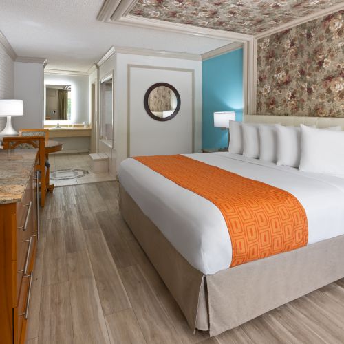 A hotel room with a large bed, orange runner, floral wallpaper accent wall, wooden furniture, a TV, desk, and a hallway leading to a mirror-lit vanity.