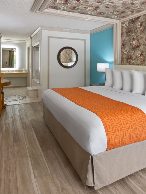 A hotel room with a large bed, orange runner, floral wallpaper accent wall, wooden furniture, a TV, desk, and a hallway leading to a mirror-lit vanity.