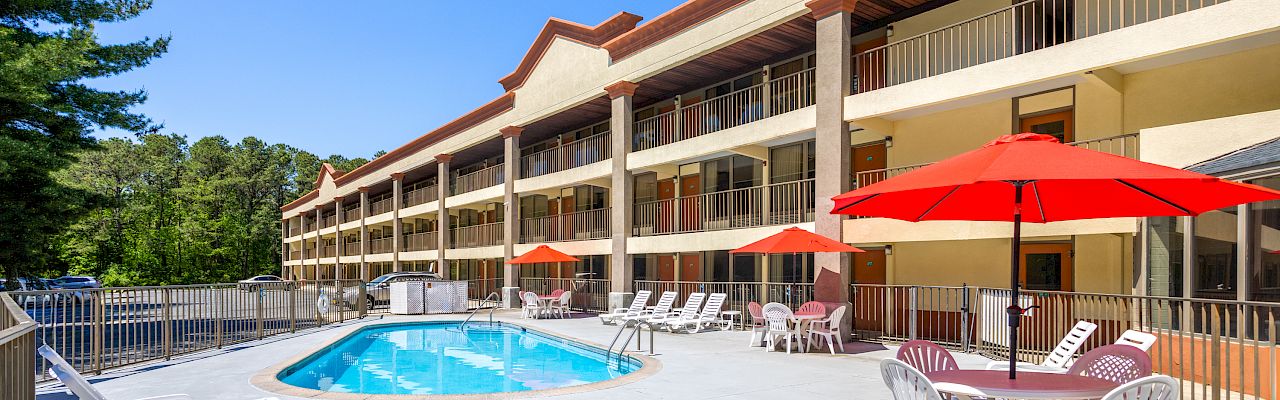 A sunny motel-style pool area with a long two-story beige building, red umbrellas, lounge chairs, and a bright blue pool.